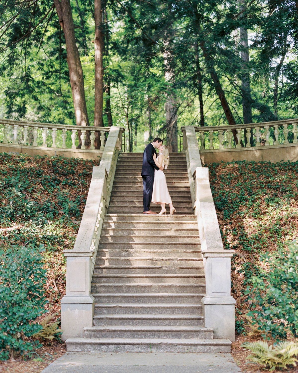 Couple kissing on stone stairs surrounded by green trees and garden foliage.