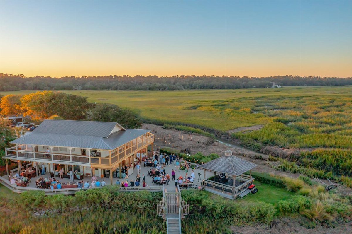 Wooden lodge and gazebo overlooking a green marshland during a sunset event.