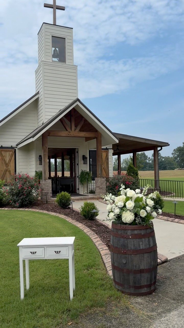 White wooden chapel with a cross, white flowers in a barrel, and a side table.