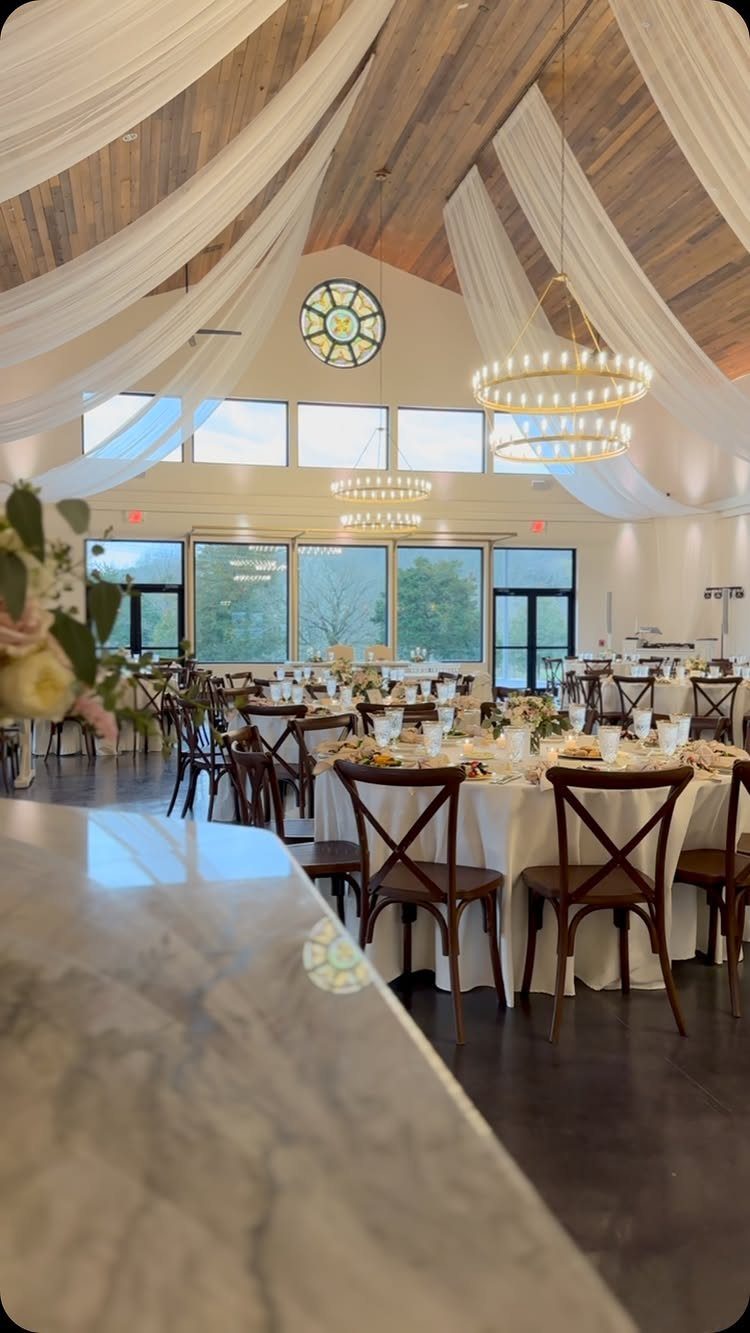 Wedding reception venue with vaulted wooden ceilings, white fabric draping, and set tables.
