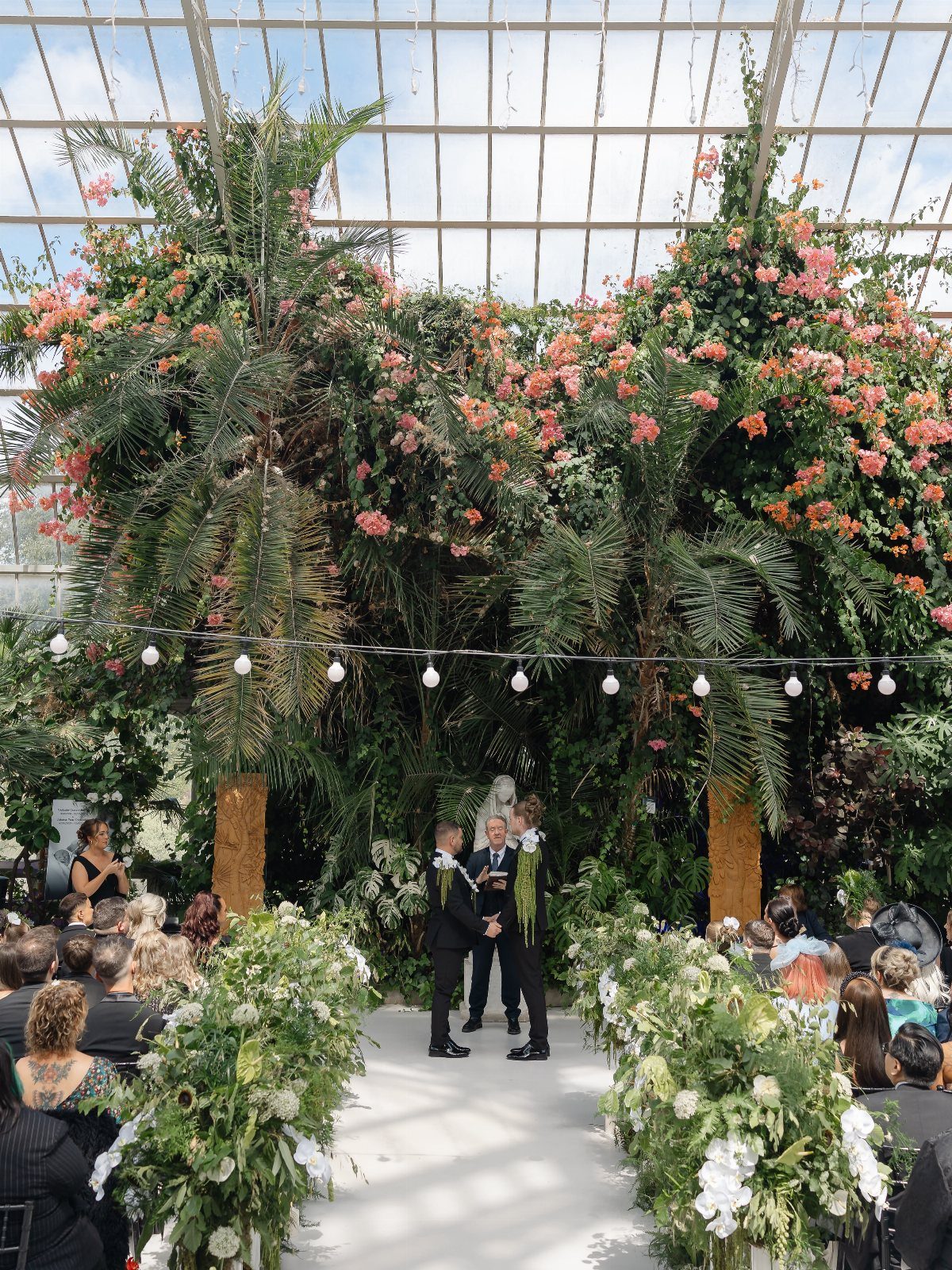 Wedding ceremony in a glass greenhouse with tropical plants and white floral arrangements.