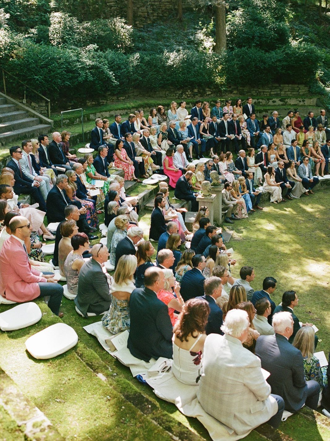 Wedding guests in formal wear seated on grassy tiers in a lush garden