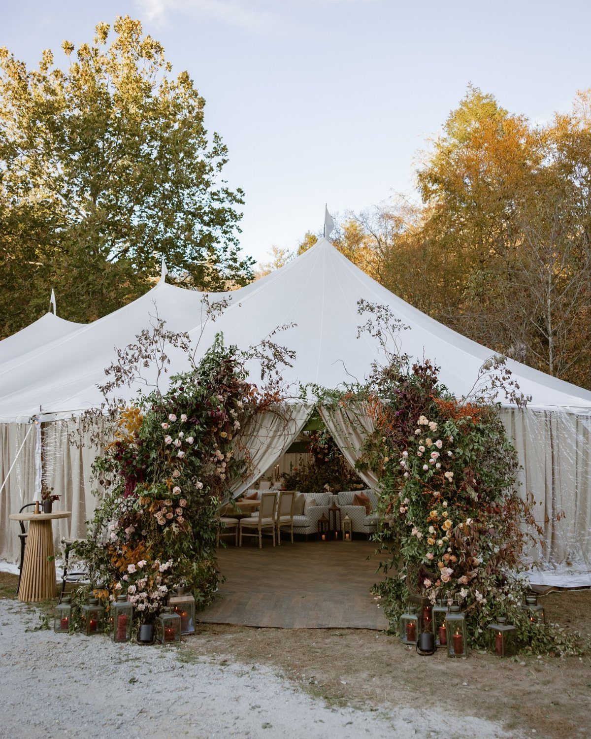 White event tent entrance framed by large autumn floral arrangements in a wooded setting