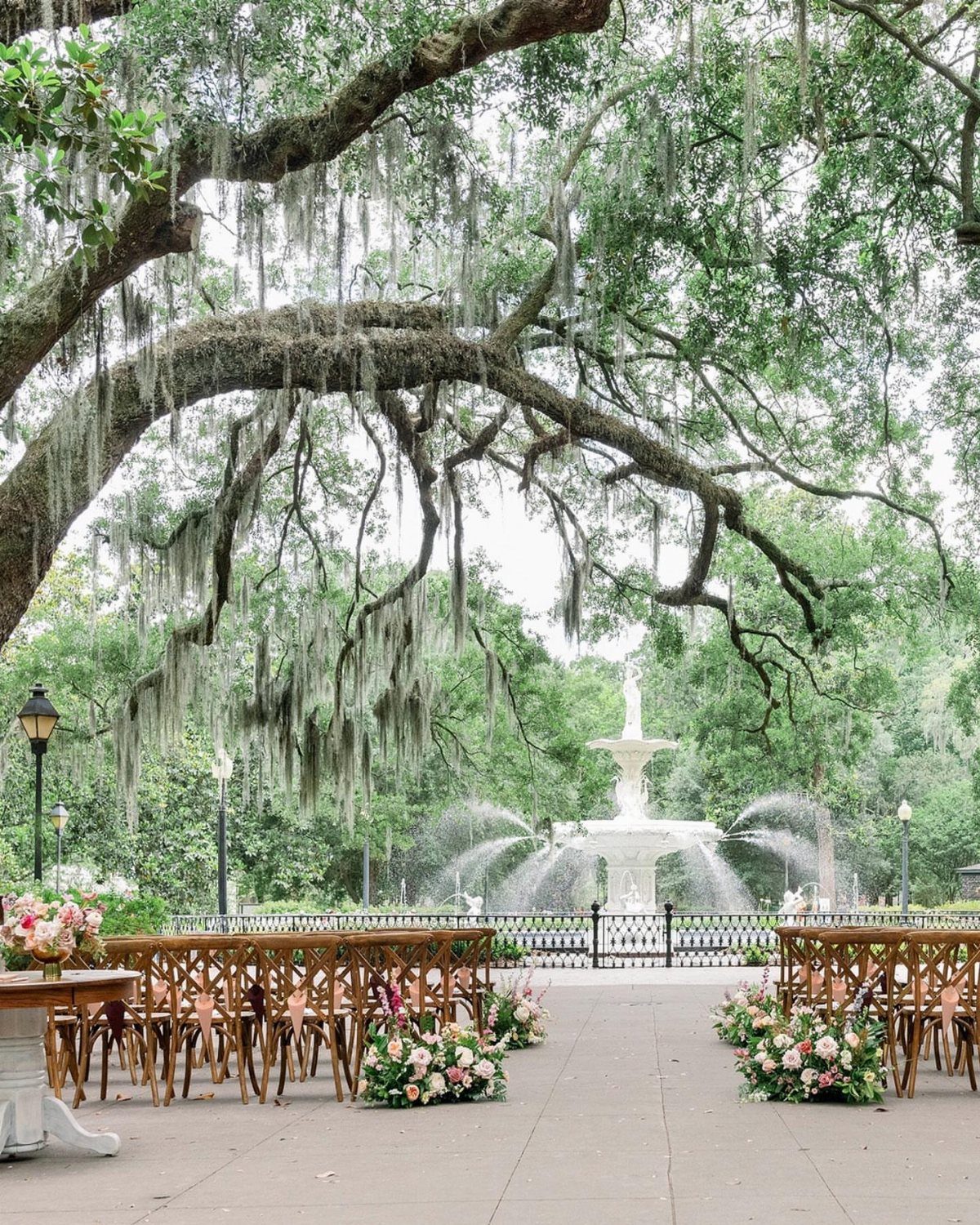 Wooden wedding chairs facing a white fountain under a mossy oak tree.