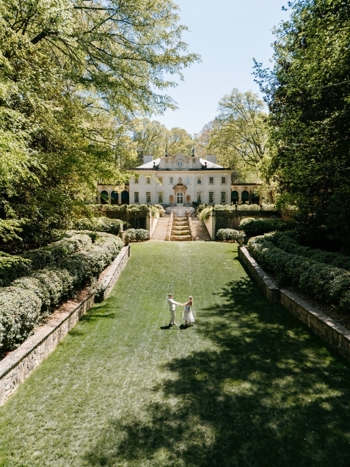 Bride and groom standing on a long lawn before a historic white mansion.