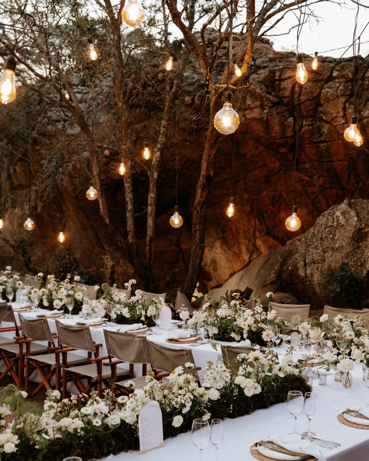 Outdoor wedding reception tables with white flowers and hanging lights against desert rocks.