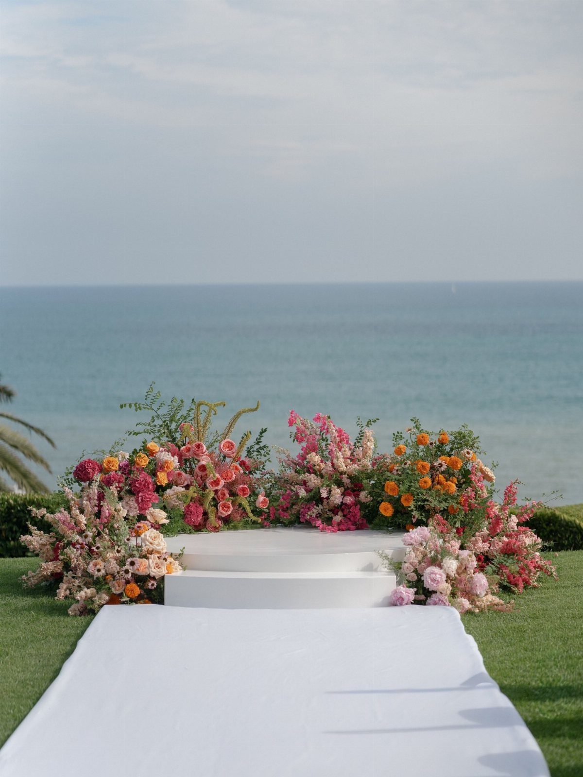 White wedding altar with colorful floral arrangements overlooking a calm blue ocean.