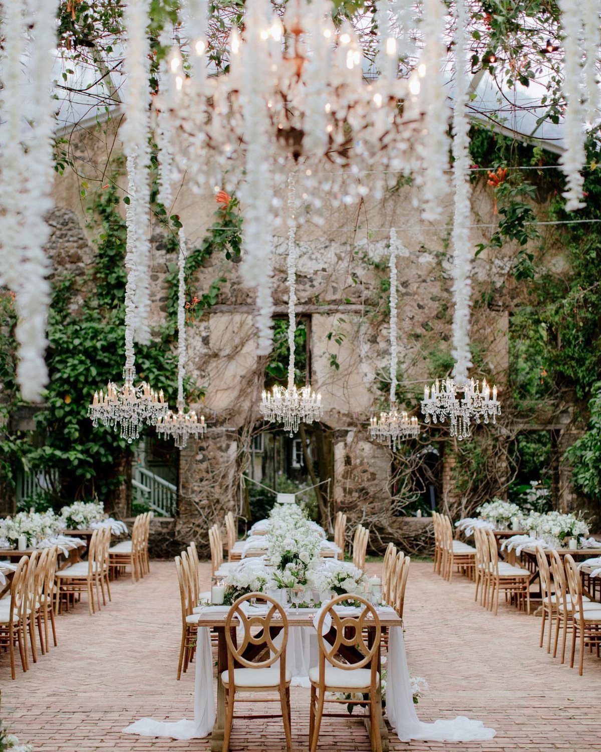 Wedding reception with gold chairs and chandeliers under white wisteria by stone walls