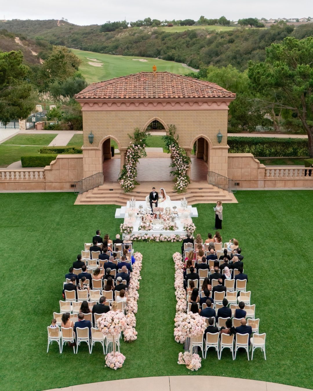Wedding ceremony at a stone pavilion with guests seated on a green lawn.