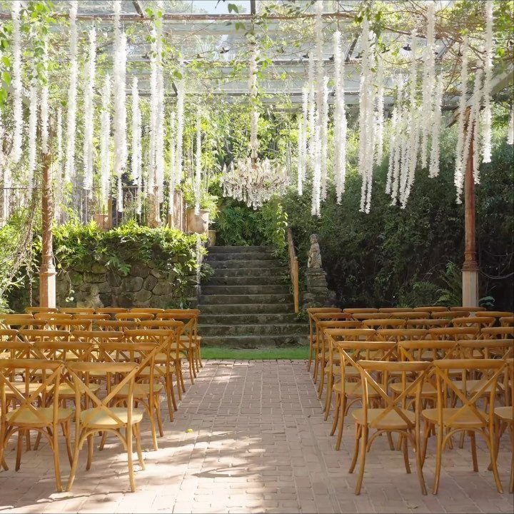 Wedding ceremony setup with wooden chairs and hanging white flowers under a glass roof.