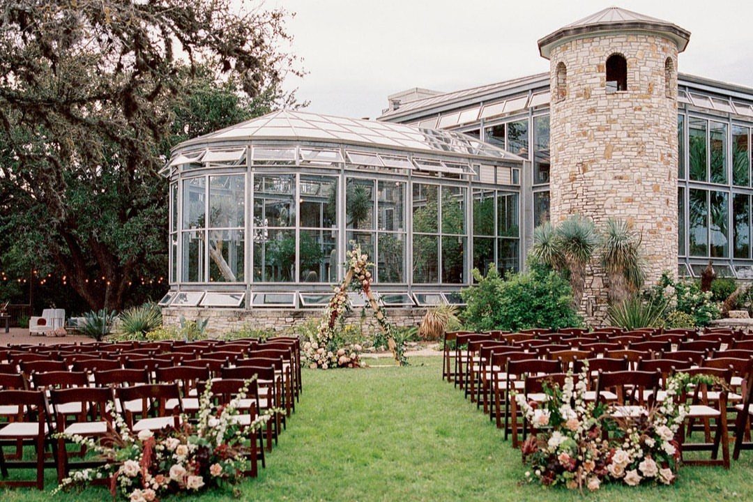 Outdoor wedding ceremony setup with wooden chairs facing a glass greenhouse and floral arch.