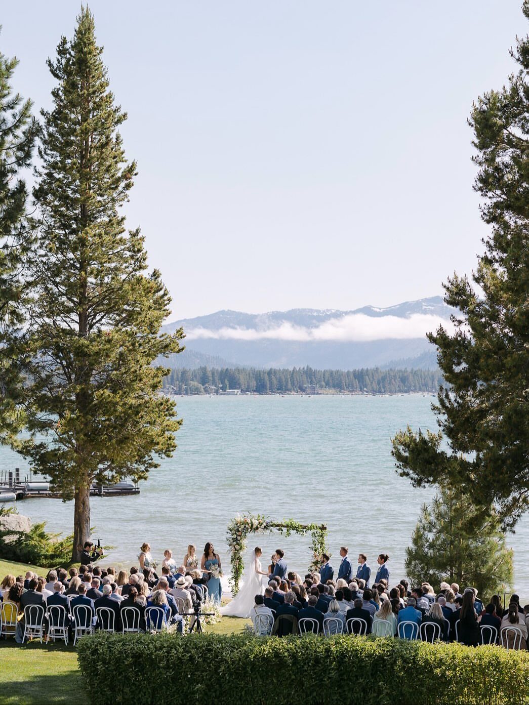 Outdoor wedding ceremony by a lake with mountain views and evergreen trees.