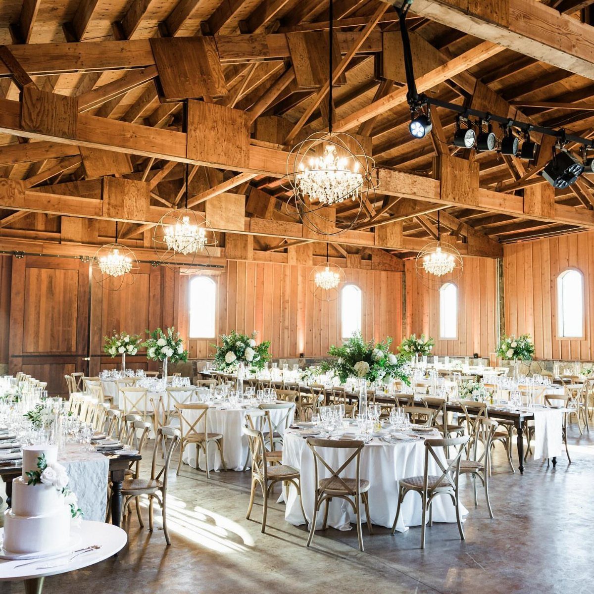 Rustic barn wedding reception with white tables, wooden chairs, and crystal chandeliers.