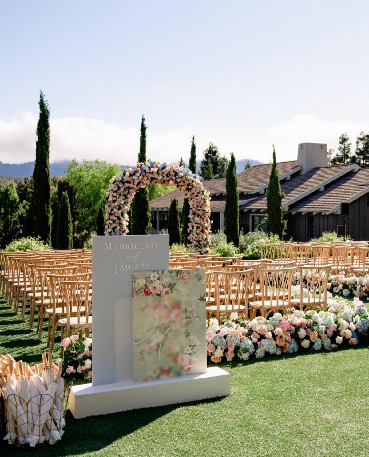 Outdoor wedding ceremony setup with wooden chairs, floral arch, and welcome sign on lawn.