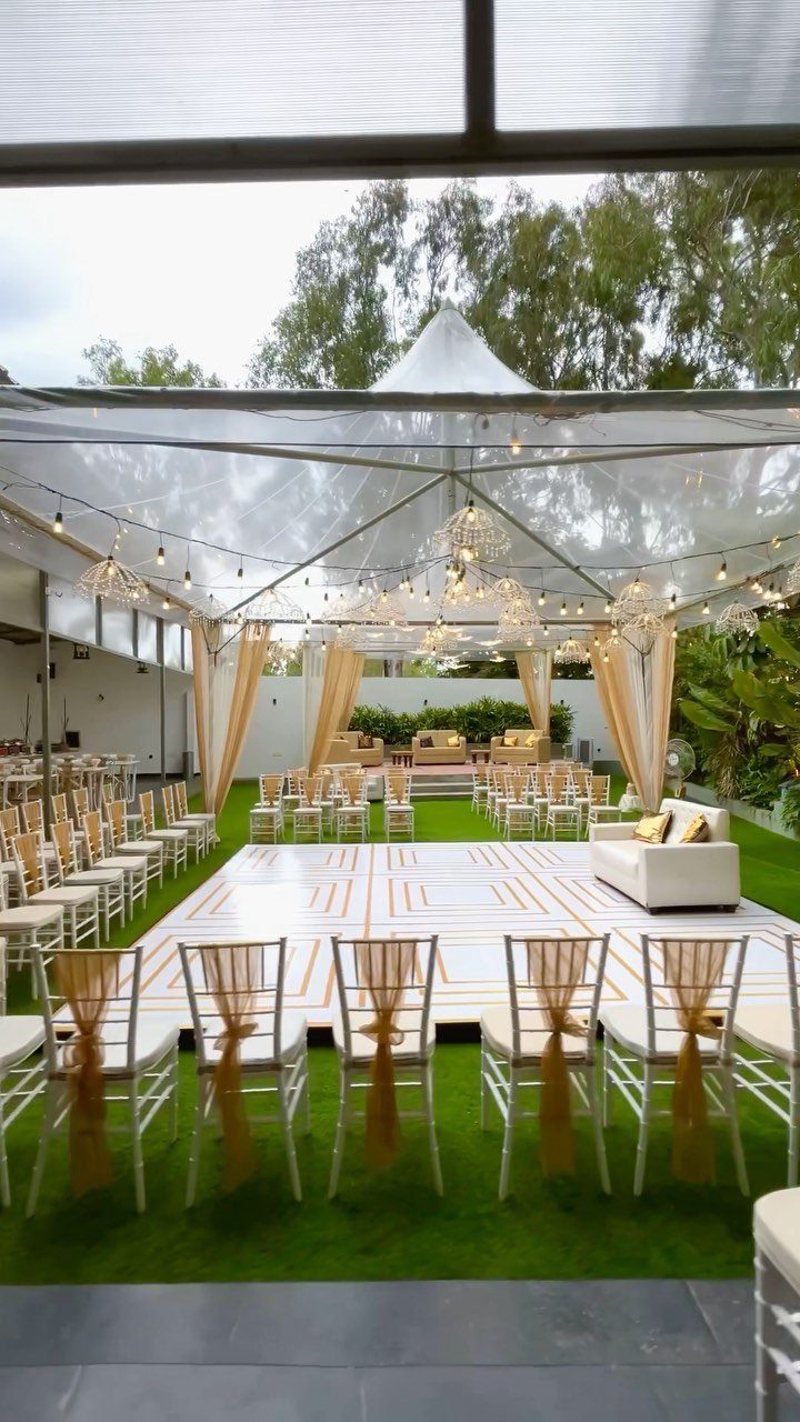 Outdoor event space with a clear tent, white dance floor, and gold chairs.