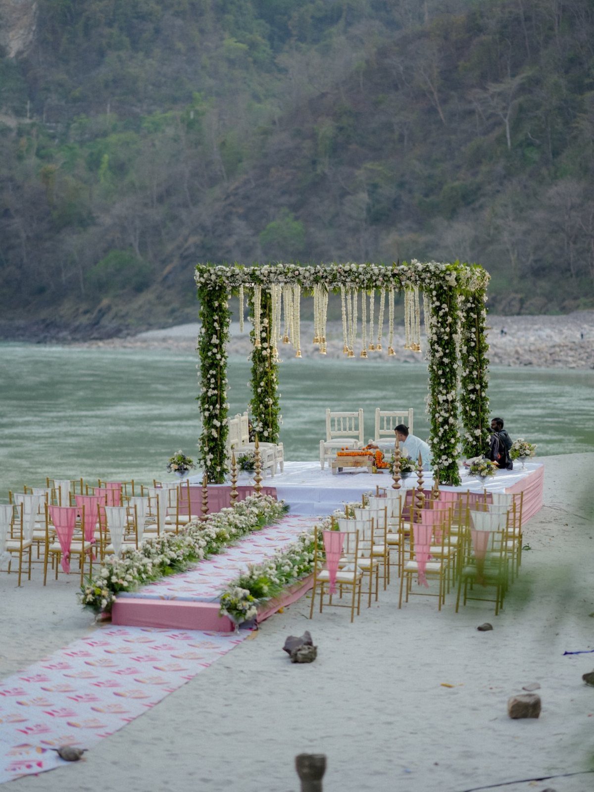 Floral wedding mandap and seating area on a sandy riverbank with mountain backdrop