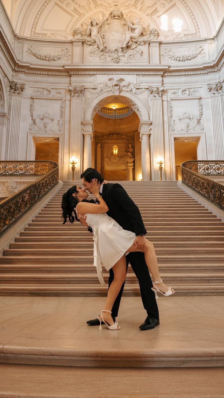 Couple in wedding attire kissing on a grand, ornate marble staircase in city hall.