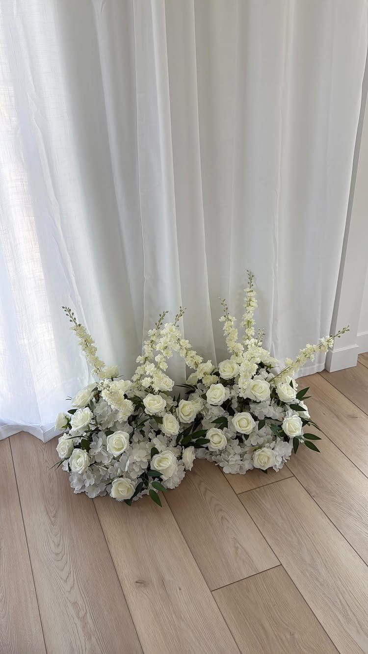 White floral arrangements on a light wood floor against white curtains.