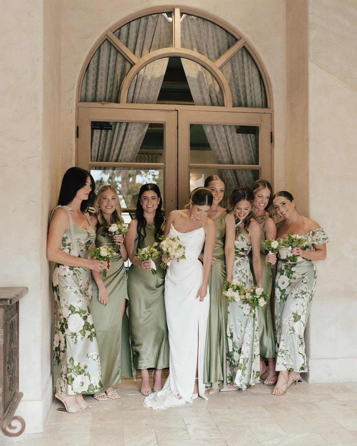 Bride and bridesmaids wearing mismatched sage green and floral dresses posing together.