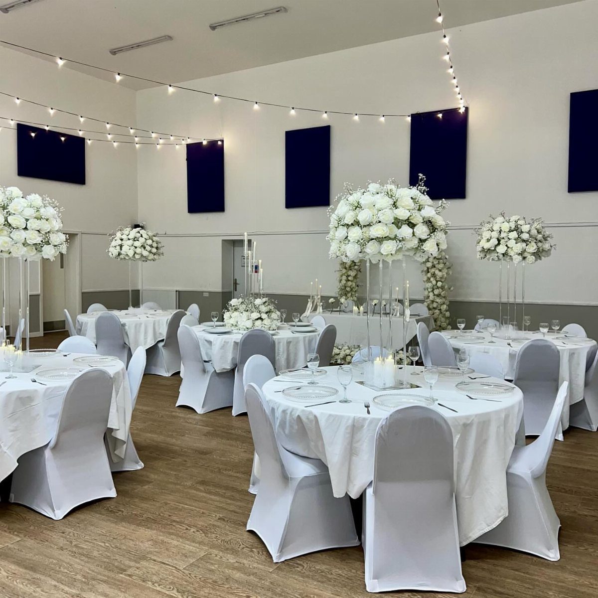 Wedding reception hall with white table linens, floral centerpieces, and overhead string lights.