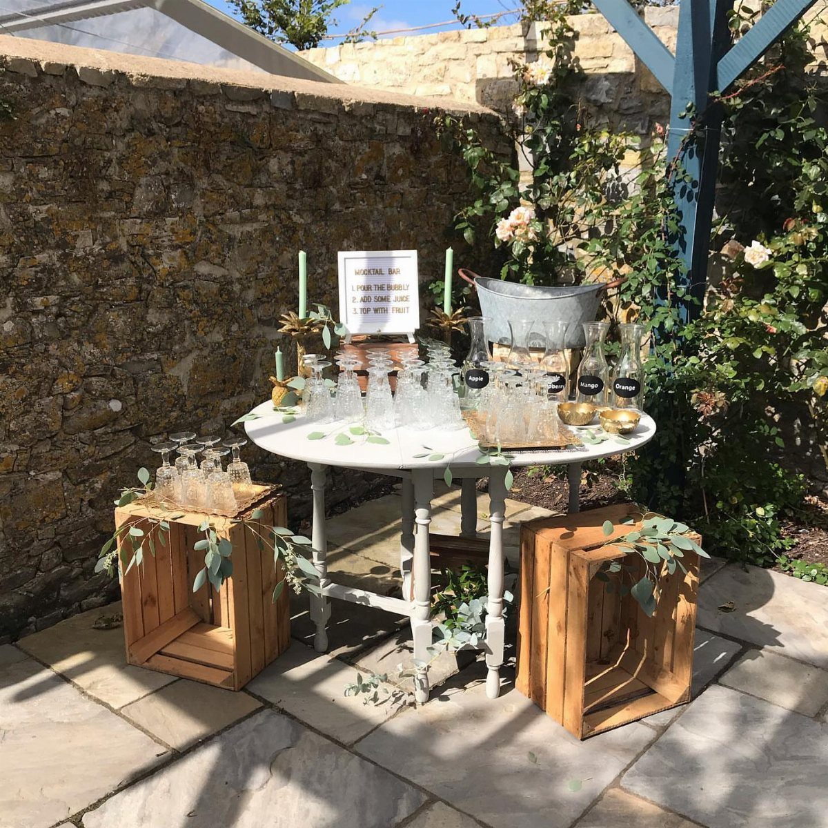 Self-serve drink station with glasses and bottles on a white table outdoors.