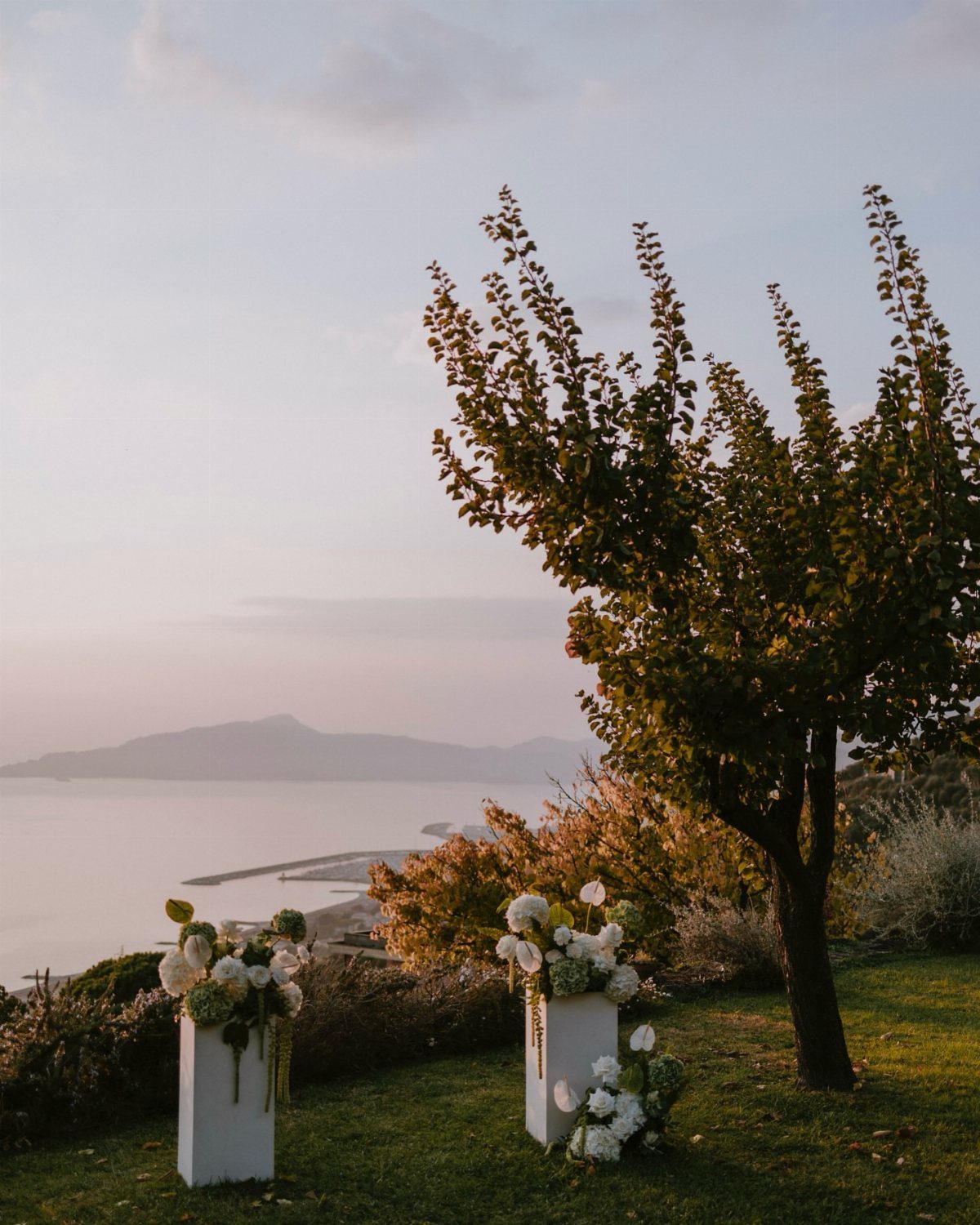 White floral arrangements on pedestals overlooking a coastal mountain landscape at sunset.