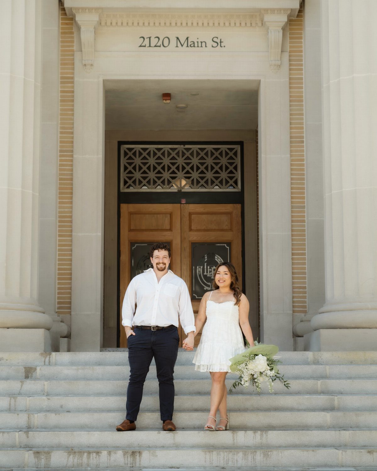 Couple in wedding attire posing on the steps of a courthouse building.