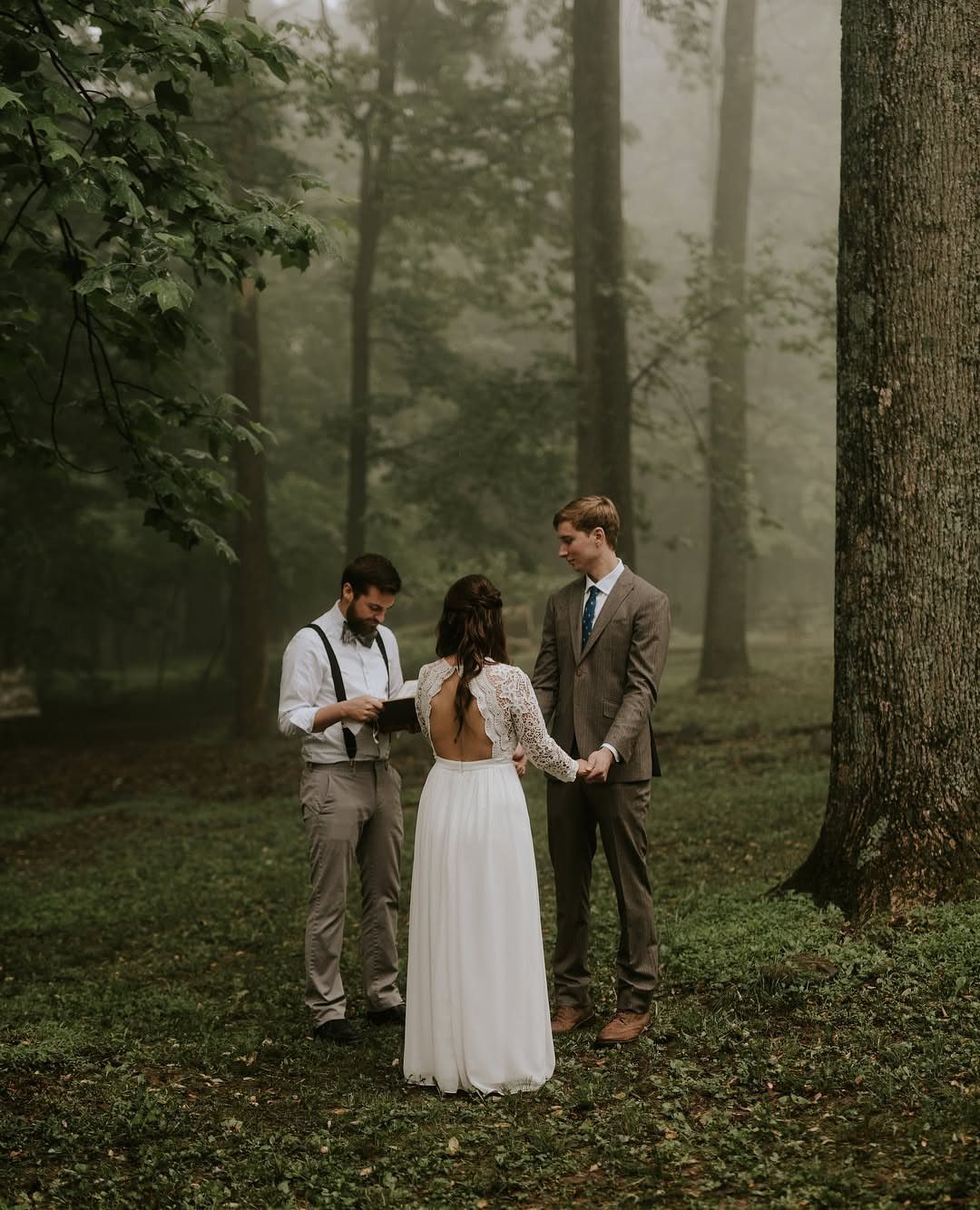 Bride and groom holding hands during a foggy forest wedding ceremony with an officiant.