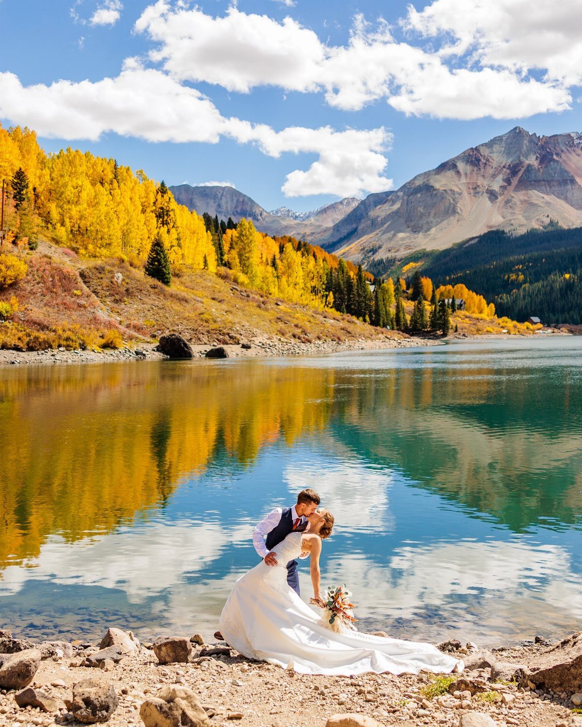 Newlywed couple kissing on a rocky shore by a mountain lake with autumn foliage.