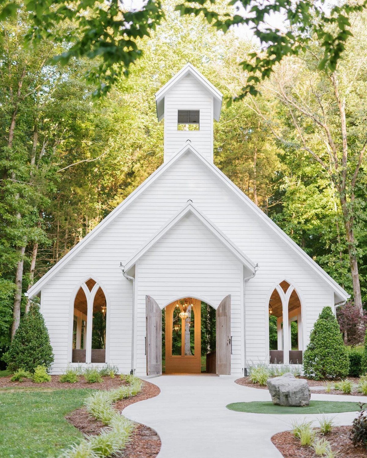 Small white wooden chapel with pointed arch windows nestled in a green forest.