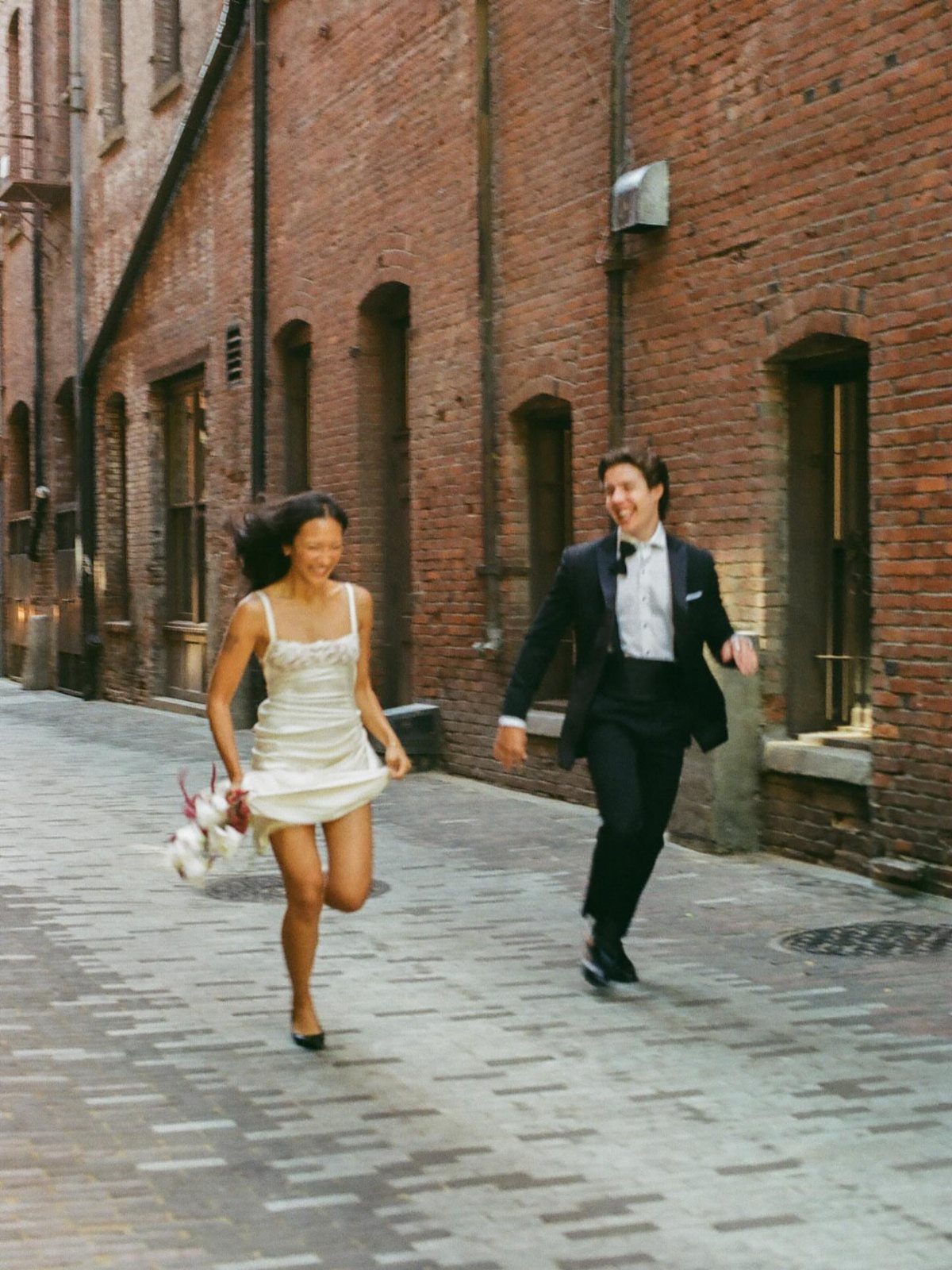 Newlywed couple in wedding attire running happily through a red brick alleyway