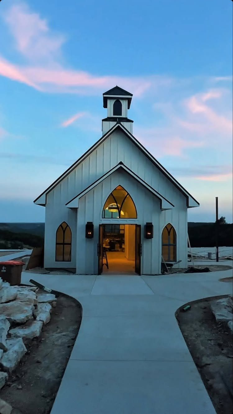 Small white wooden chapel with glowing interior under a twilight sky.