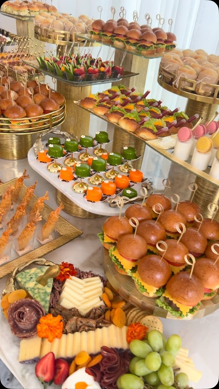 Catering spread with mini sliders, charcuterie, and colorful appetizers on gold stands.
