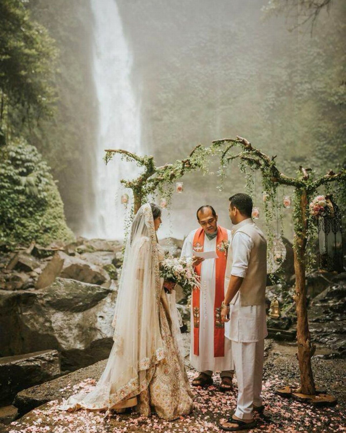 Bride and groom standing under a floral arch with a waterfall background.