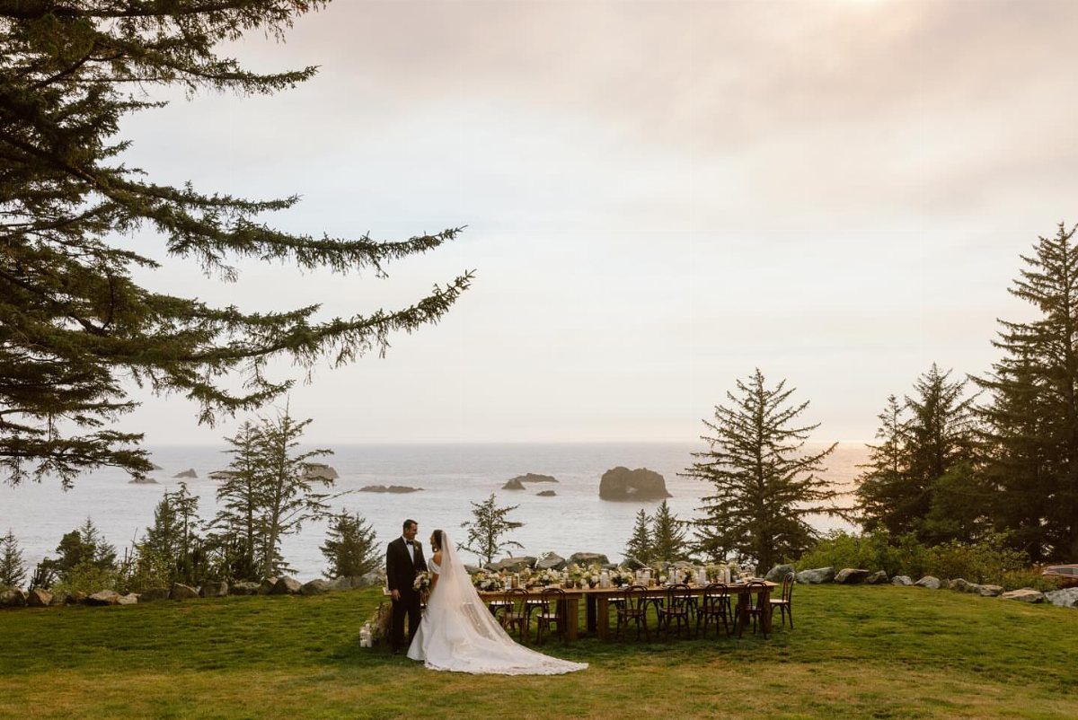 Bride and groom standing by a long wedding table overlooking a rocky coastline.