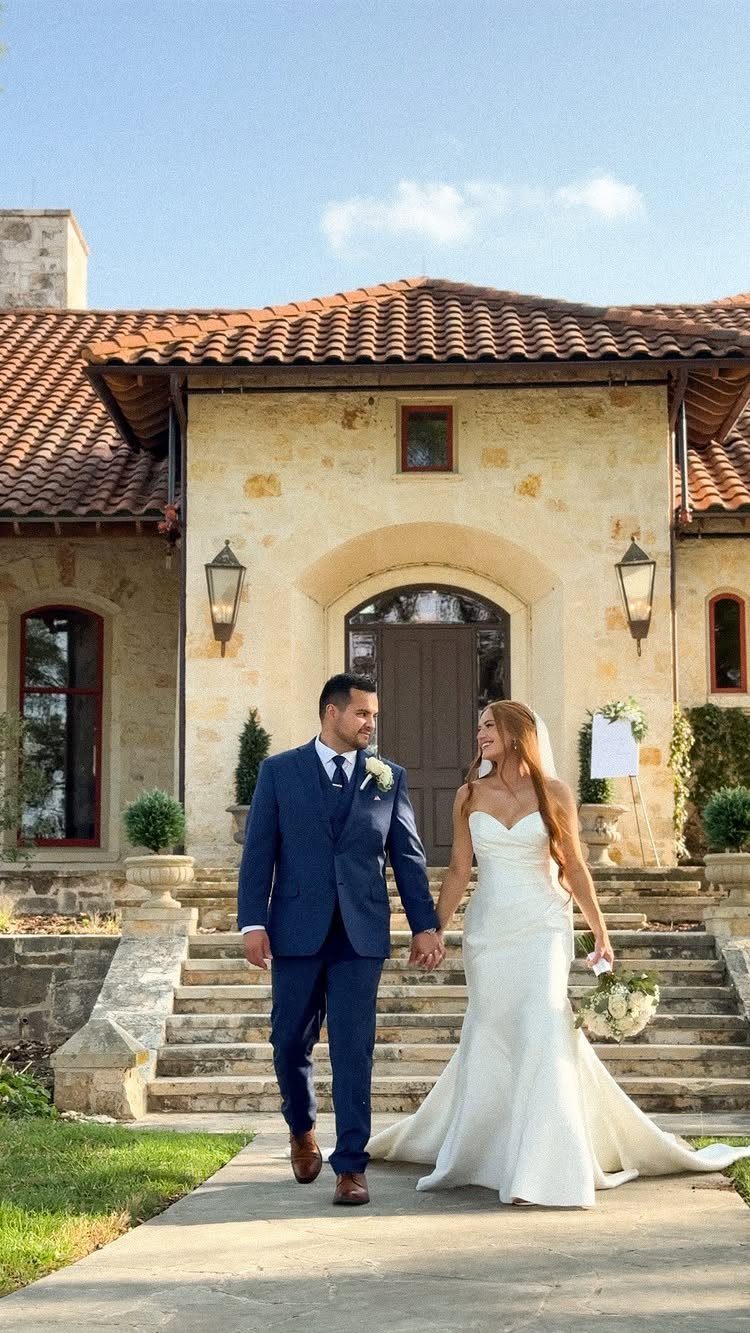 Bride and groom walking hand in hand in front of a stone Tuscan villa.