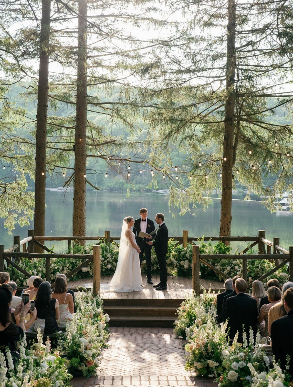 Wedding ceremony on a wooden deck overlooking a lake surrounded by tall pine trees.