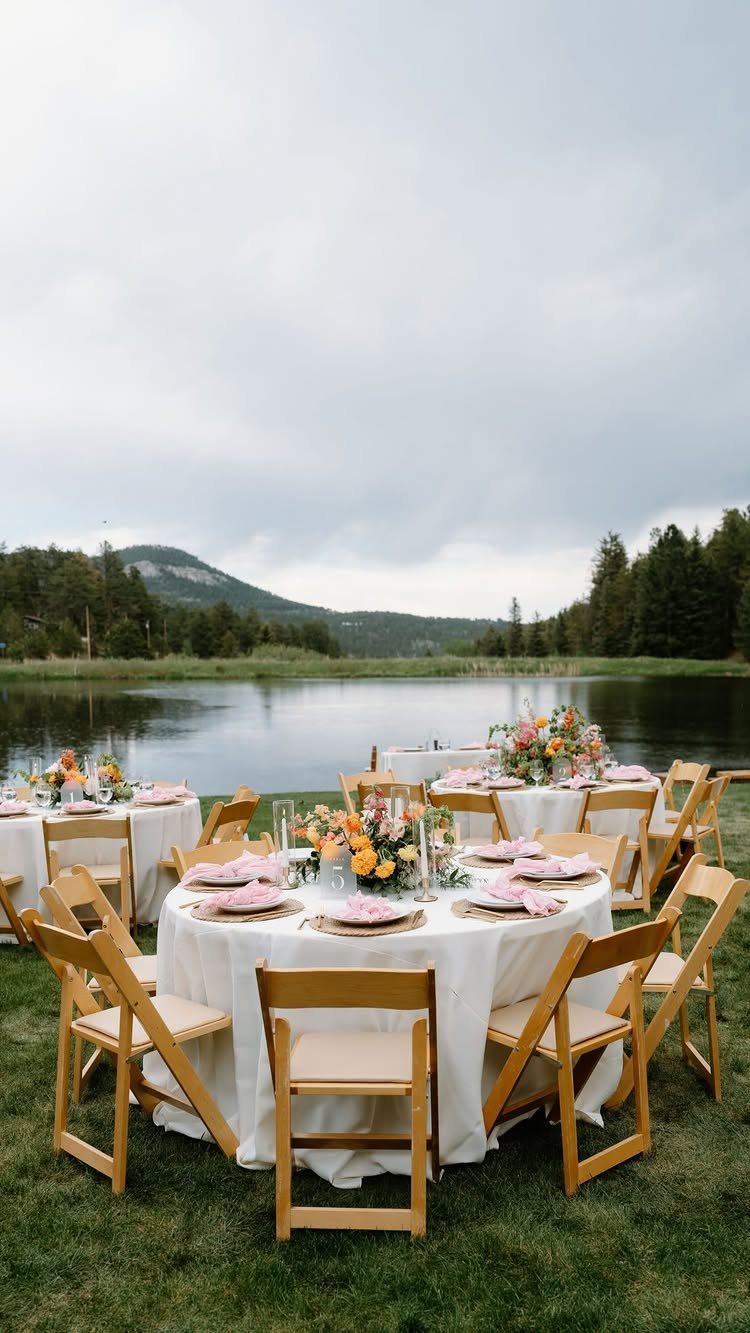 Round wedding tables with white linens and flowers by a lake and mountains.