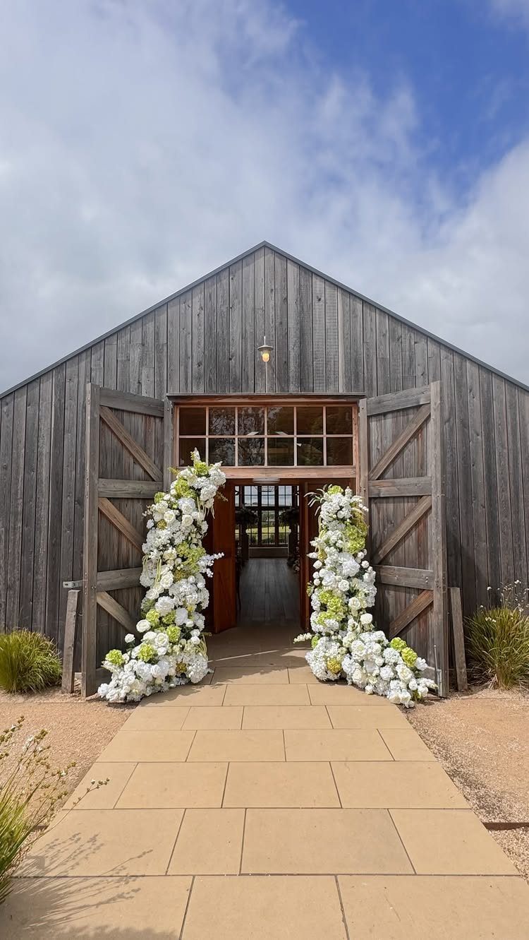 Grey wooden barn entrance decorated with white and green floral arrangements and stone path