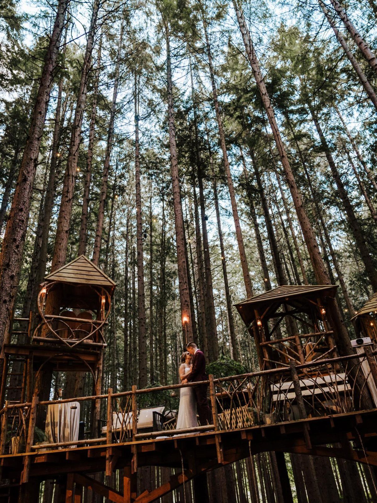 Bride and groom embracing on a wooden bridge between forest treehouses.