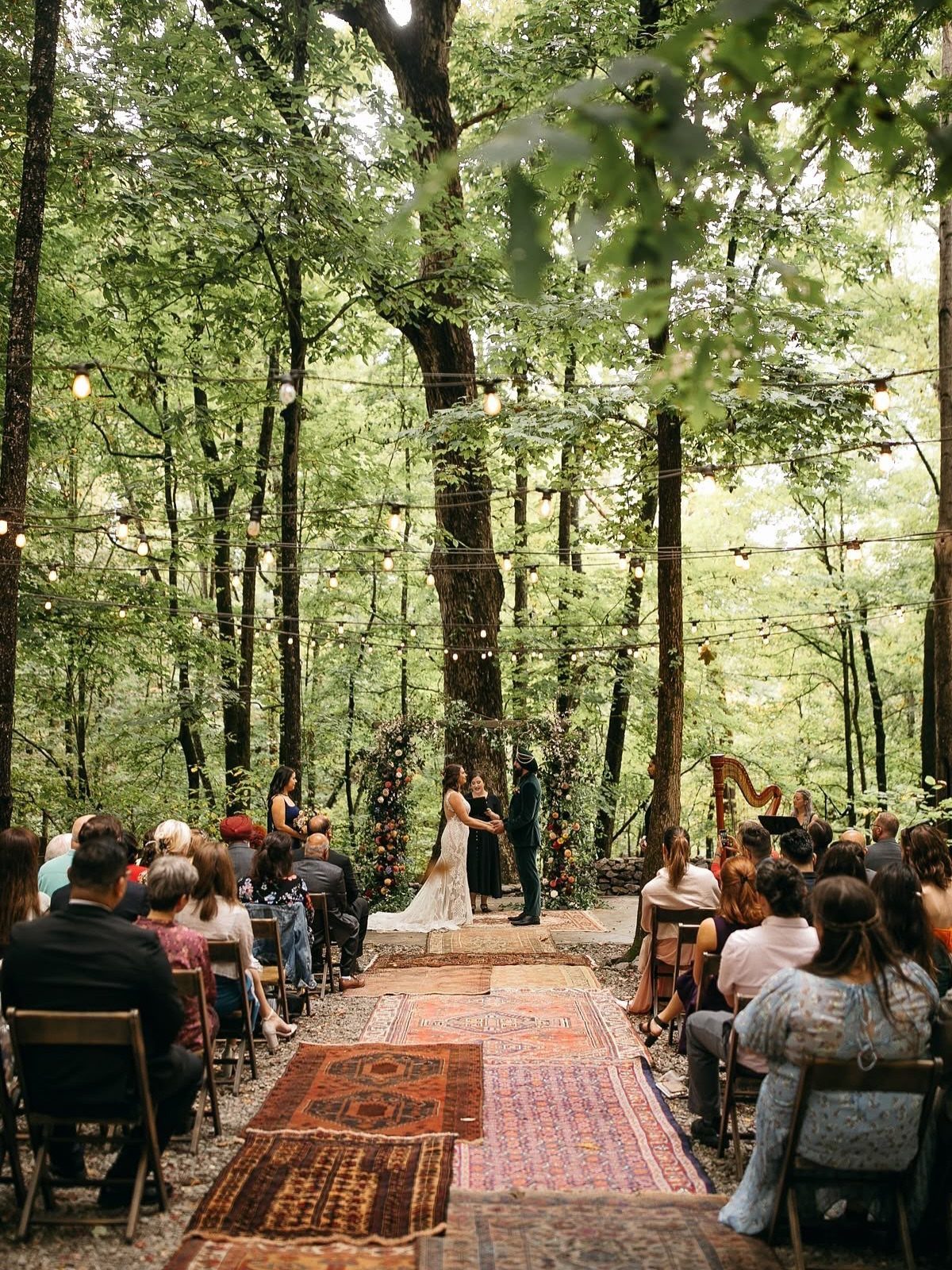 Wedding ceremony in a forest with string lights and patterned rugs on the ground.