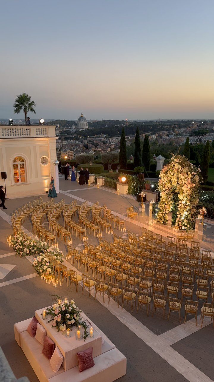 Outdoor wedding ceremony setup on a terrace overlooking the Rome city skyline at dusk.