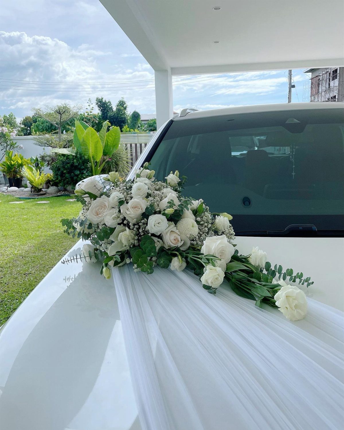 White roses and greenery draped across a white car hood with sheer fabric.