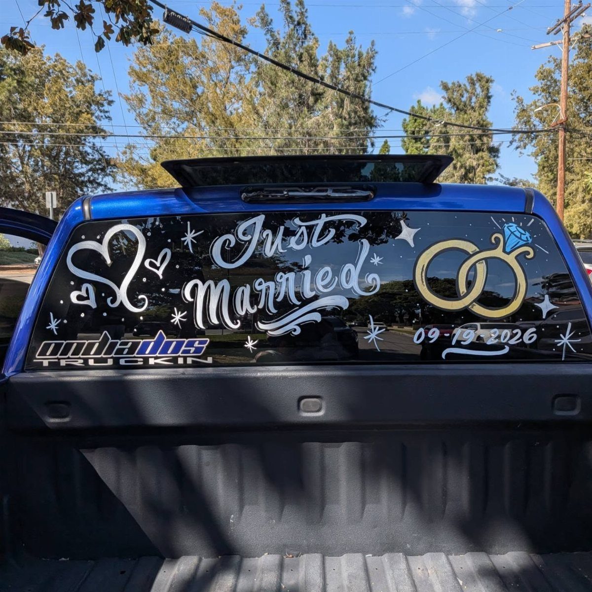 Blue pickup truck rear window painted with Just Married text and wedding rings.