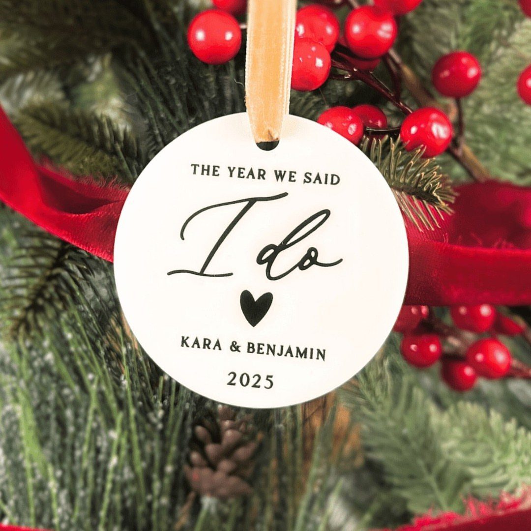 White ceramic Christmas ornament with wedding text hanging on a festive evergreen tree.