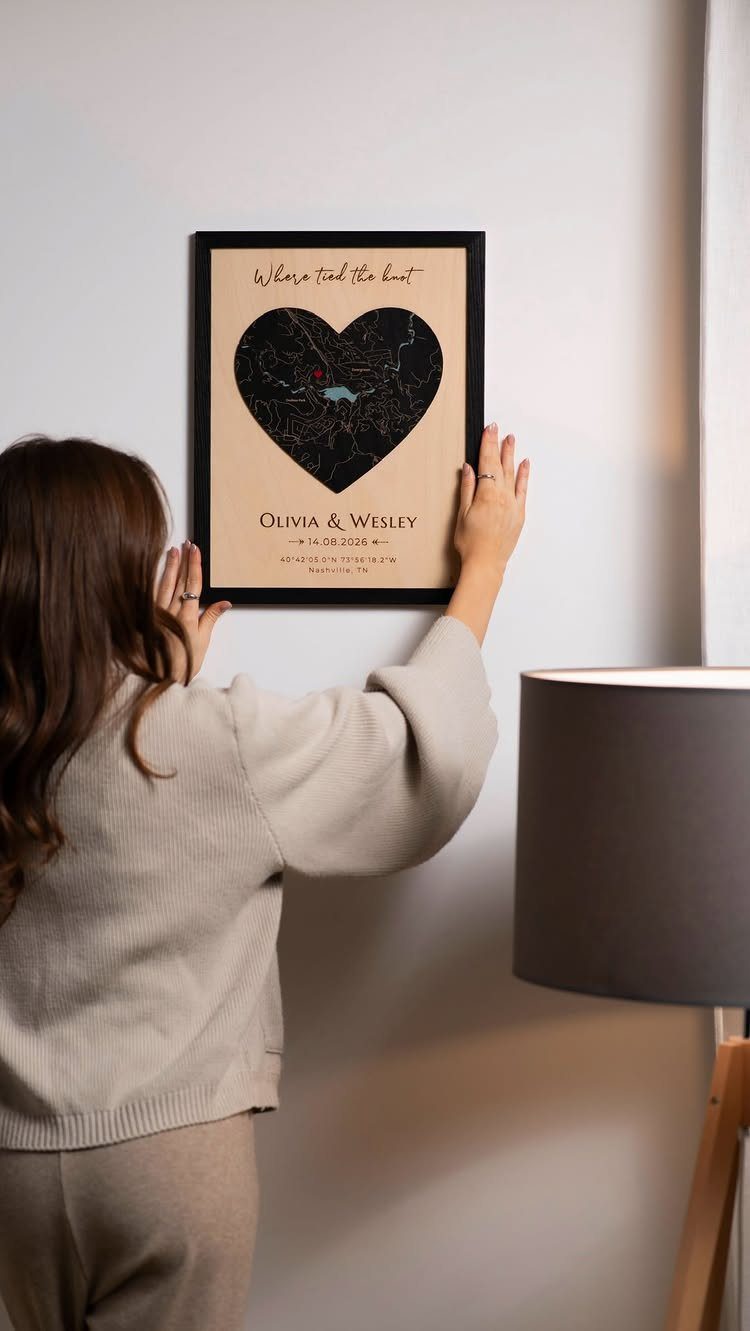 Woman hanging a heart-shaped personalized map art frame on a white wall.