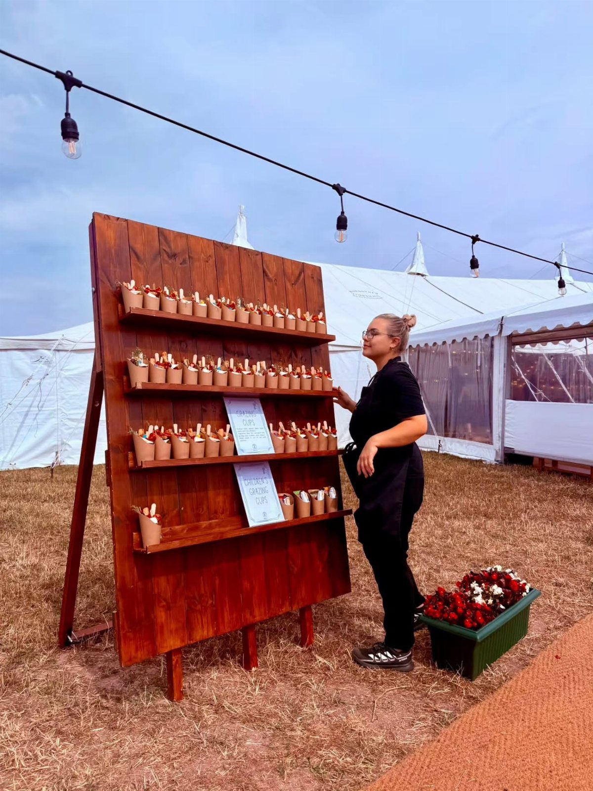 Woman standing by a wooden dessert display stand at an outdoor event.