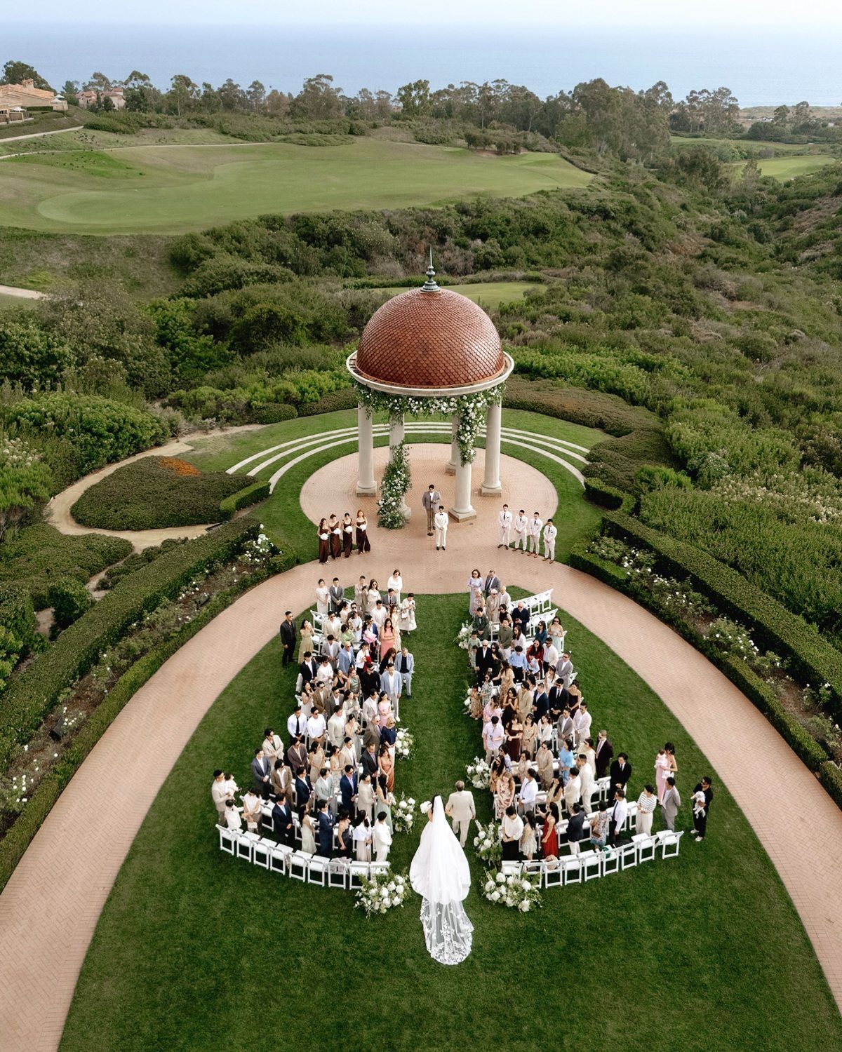 Outdoor wedding ceremony with guests seated before a red-domed gazebo overlooking the ocean.
