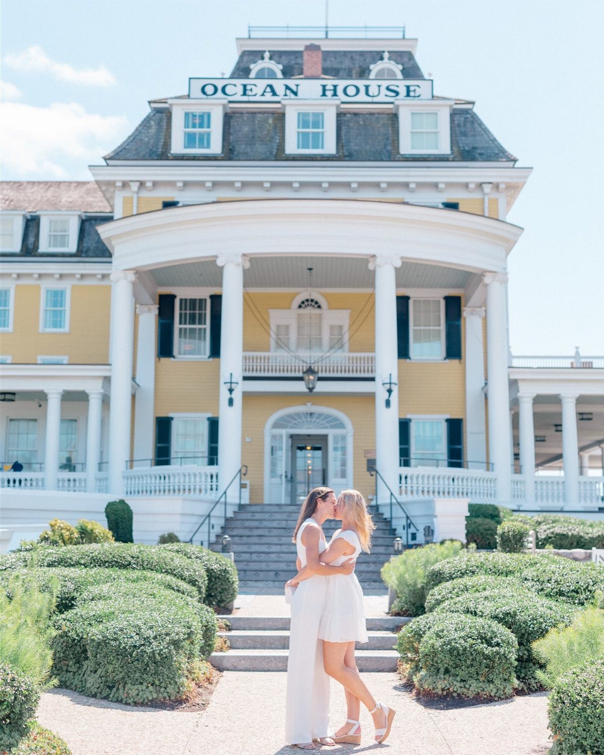 Two women in white dresses kissing before the yellow Ocean House hotel facade.
