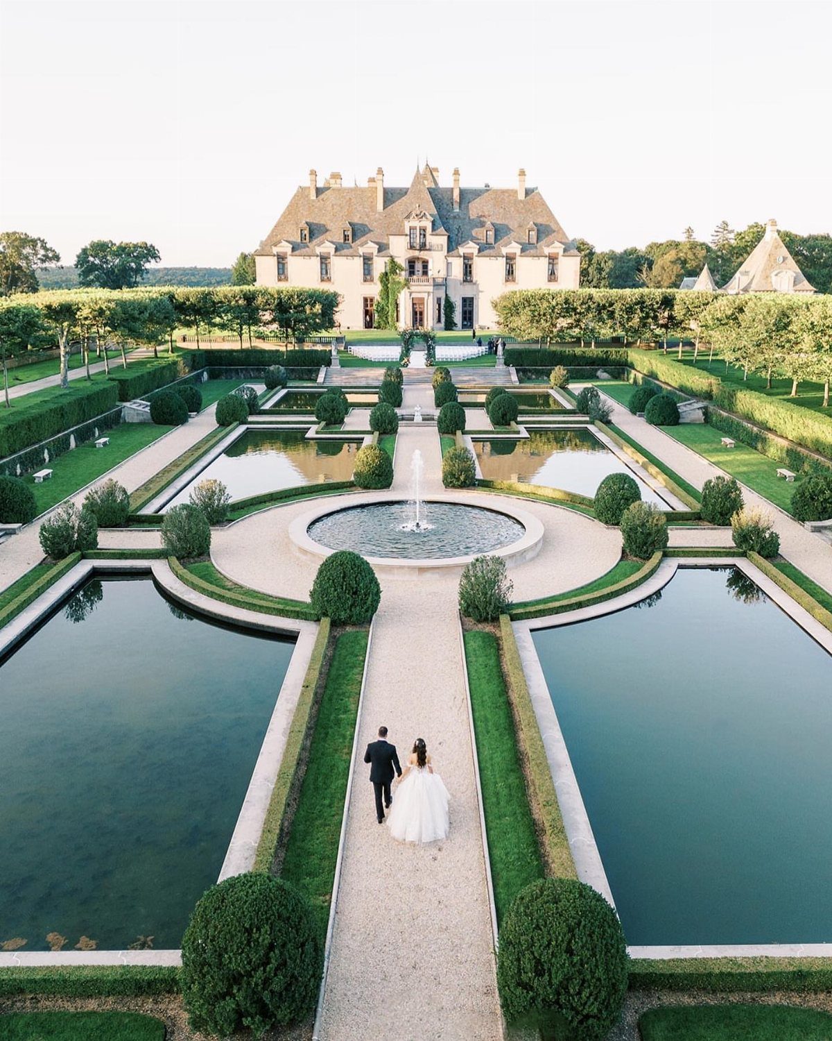 Bride and groom walking toward a grand stone castle through formal garden pools.